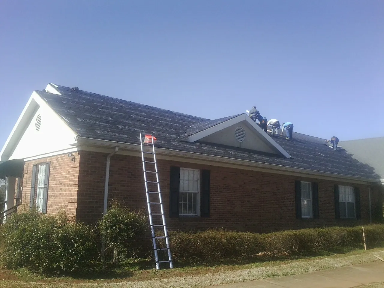 Crew tearing shingles off a brick ranch roof with a ladder set — Metal Roof Repair work by our crew in Nerstrand
