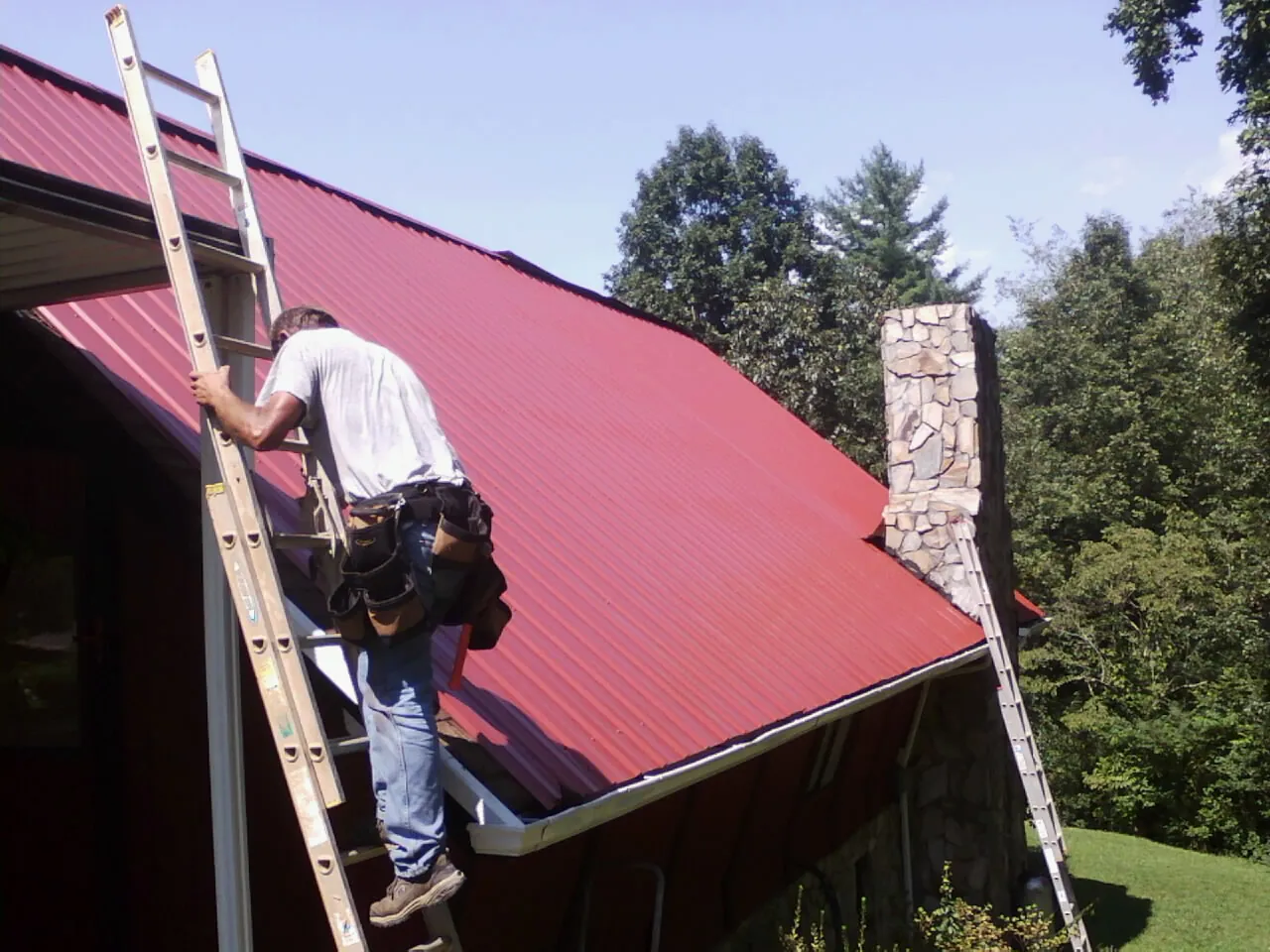 Roofer on a ladder beside a red metal roof and stone chimney — Soffit & Fascia Repair by Roofing Brothers in Nerstrand